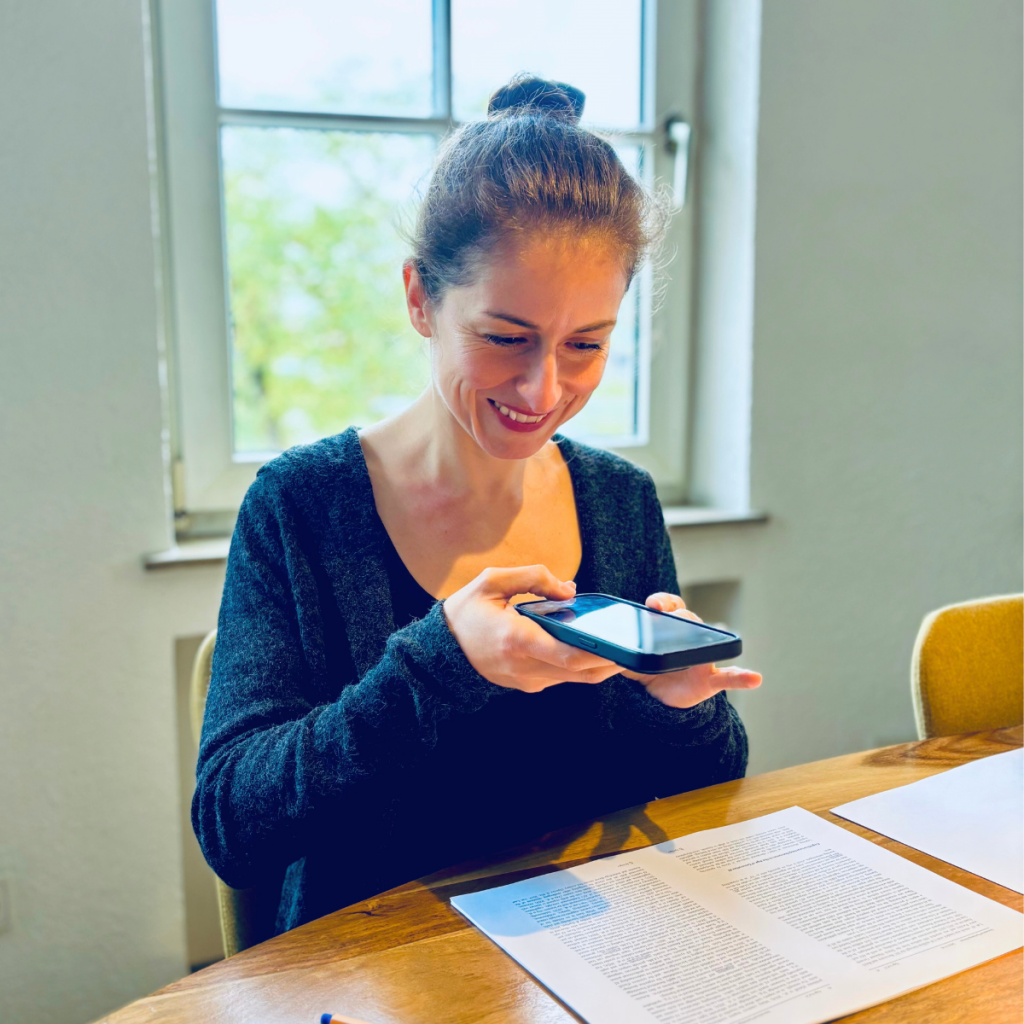 Jacqueline Sammet sitzt am Tisch und hält ein Smartphone über eine DIN-A-4 Seite